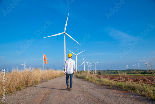 Engineer wearing uniform ,helmet hold document inspection work in wind turbine farms rotation to generate electricity energy. Green ecological power energy generation wind sustainable energy concept.