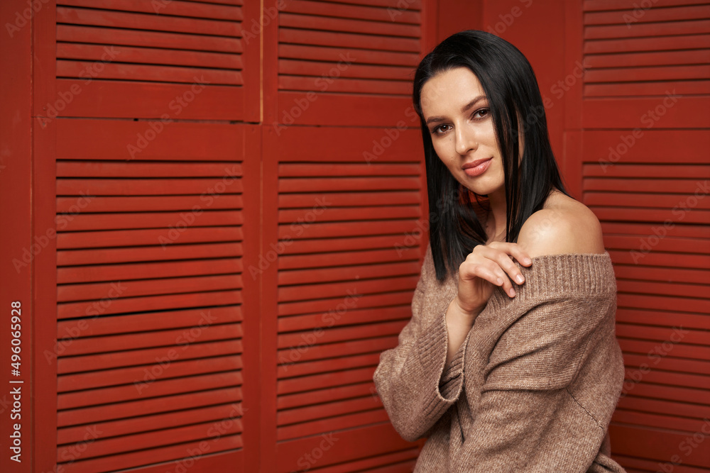 beautiful woman with dark hair in cozy clothes with a bare shoulder against the background of a wooden red wall