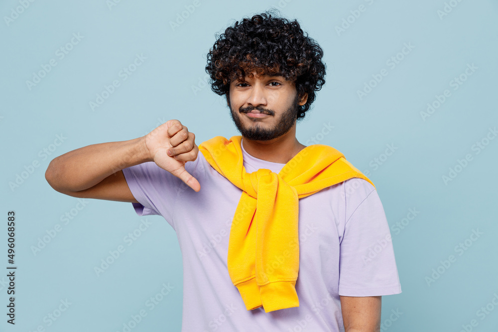 Unhappy distempered sad unnerved aggrieved young bearded Indian man 20s years old wears white t-shirt showing thumb down dislike gesture isolated on plain pastel light blue background studio portrait. Unhappy distempered sad unnerved aggrieved young bearded Indian man 20s years old wears white t-shirt showing thumb down dislike gesture isolated on plain pastel light blue background studio portrait.