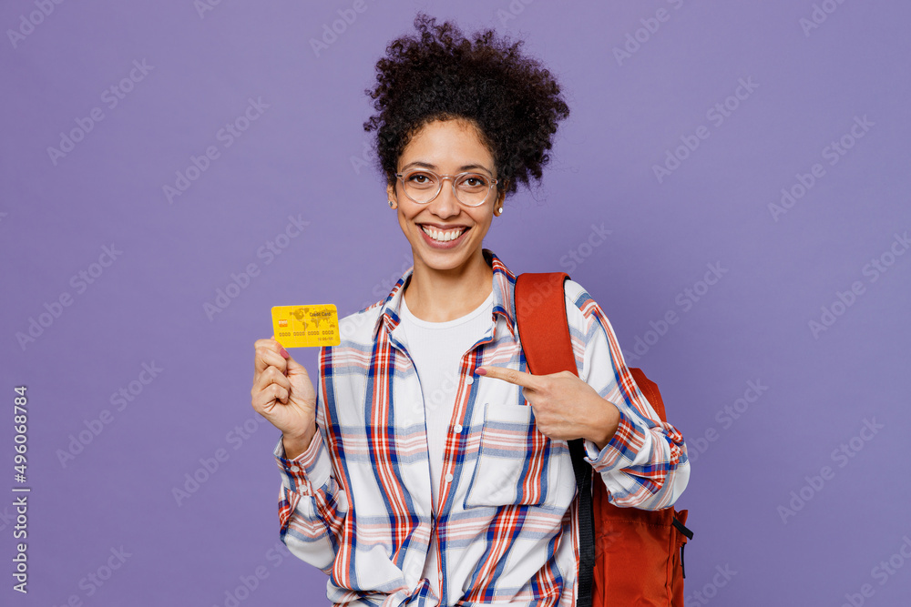Young happy girl woman of African American ethnicity teen student in shirt backpack hold books point finger on credit bank card isolated on plain purple background. Education in high school concept.