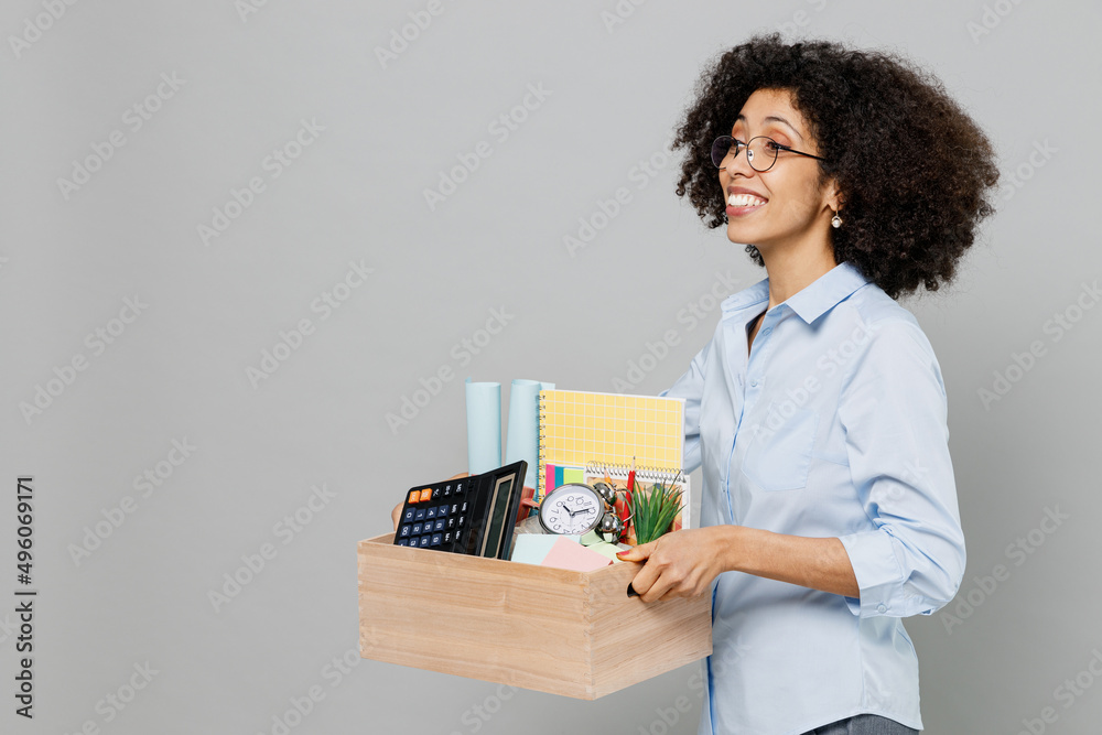 Side view young fun optimistic employee business corporate lawyer woman of African American ethnicity in classic formal shirt work in office hold cardboard box with stuff isolated on grey background. Side view young fun optimistic employee business corporate lawyer woman of African American ethnicity in classic formal shirt work in office hold cardboard box with stuff isolated on grey background.
