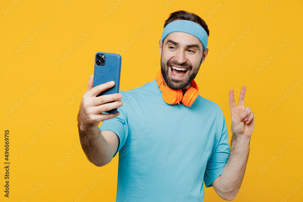 Young fitness trainer instructor sporty man sportsman in headband blue t-shirt doing selfie shot on mobile cell phone show v-sign isolated on plain yellow background. Workout sport motivation concept. Young fitness trainer instructor sporty man sportsman in headband blue t-shirt doing selfie shot on mobile cell phone show v-sign isolated on plain yellow background. Workout sport motivation concept.