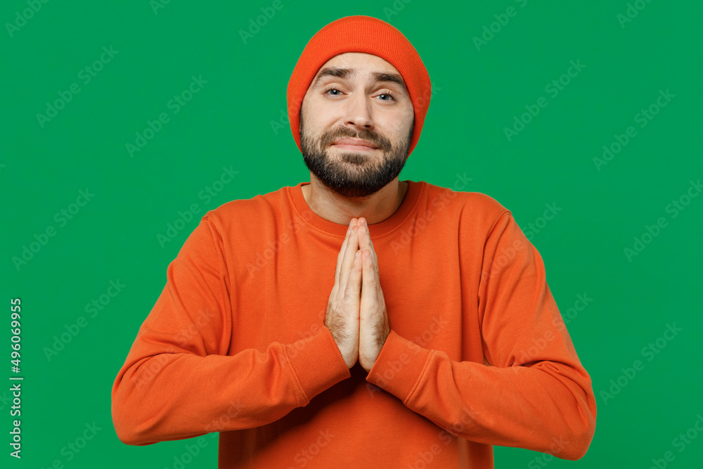 Young smiling happy caucasian man 20s in orange sweatshirt hat hold hands folded in prayer gesture begging about something isolated on plain green background studio portrait. People lifestyle concept. Young smiling happy caucasian man 20s in orange sweatshirt hat hold hands folded in prayer gesture begging about something isolated on plain green background studio portrait. People lifestyle concept.