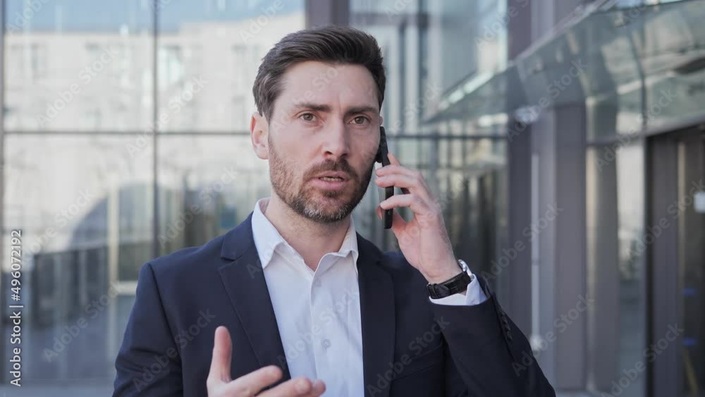 Worried nervous businessman dressed in a classic suit talking on a cell phone outdoors. Handsome angry man having bad news about job questions gesturing with hands shows his displeasure near office. Worried nervous businessman dressed in a classic suit talking on a cell phone outdoors. Handsome angry man having bad news about job questions gesturing with hands shows his displeasure near office.