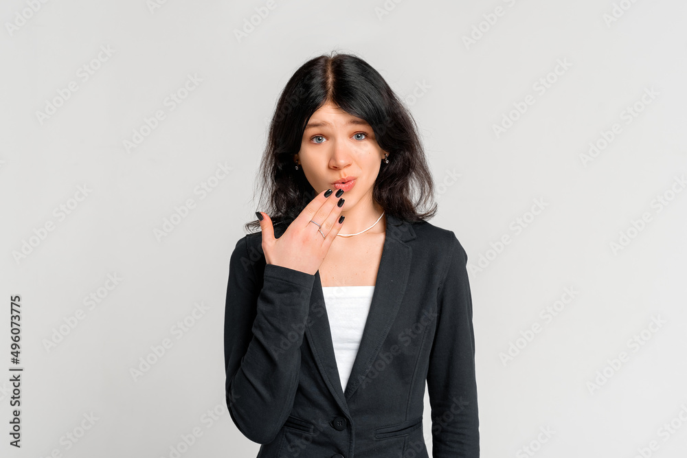 Embarrassed young dark-haired girl use hand cover mouth for not commenting and looking insecure at camera, standing in black blazer over grey background Embarrassed young dark-haired girl use hand cover mouth for not commenting and looking insecure at camera, standing in black blazer over grey background