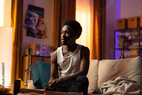 A man with an afro sits concentrated in the living room on the couch and plays a video game on the console, holding rain in his hands and eats take-out pizzas