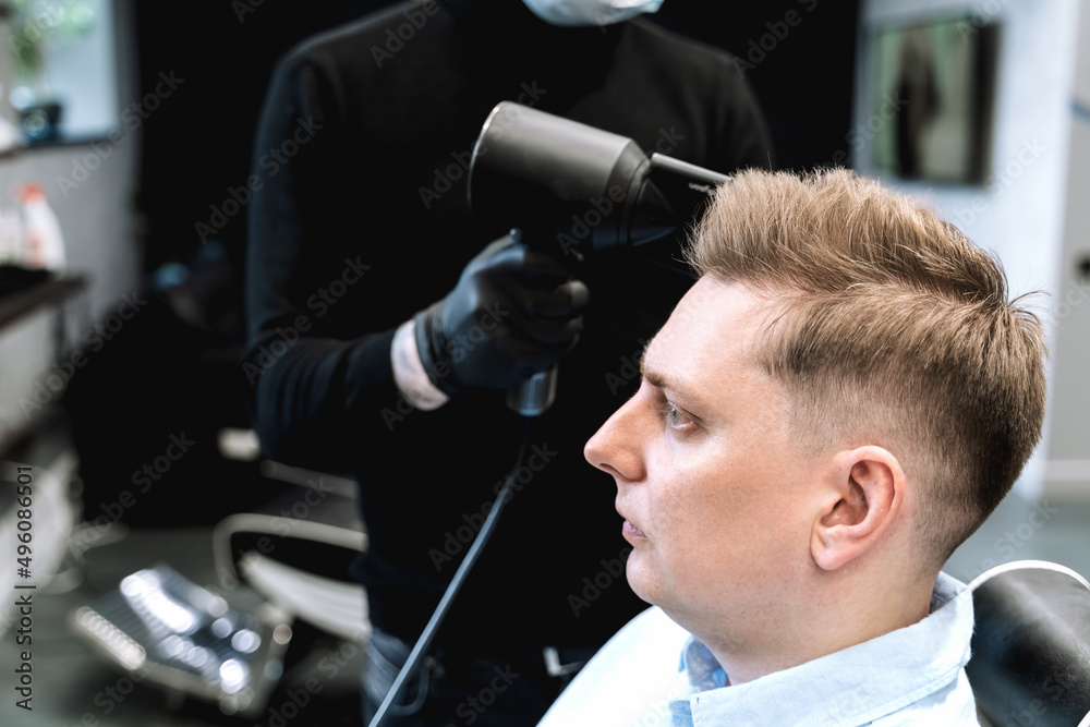 Fototapeta premium Visit to barbershop. Stylish man makes fashionable haircut. Barber,hairdresser,stylist with scissors, comb cutting,dryer.Combing hair.Client,customer sits in dark men's beauty salon covered with cape