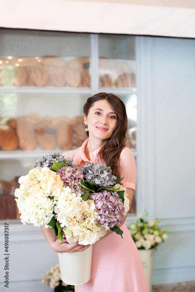 Happy woman with flowers hydrangeas in the city. Portrait of a young smiling girl wearing a pink dress holding a bouquet of flowers, posing in European street near a coffee shop. French style. Happy woman with flowers hydrangeas in the city. Portrait of a young smiling girl wearing a pink dress holding a bouquet of flowers, posing in European street near a coffee shop. French style.