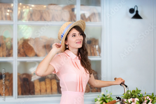 Beautiful girl with long curly hair in dress and straw hat standing with bicycle on background of street cafe. Girl ride to work by bike. Eco transport - bicycle. Young woman with a bouquet of flowers