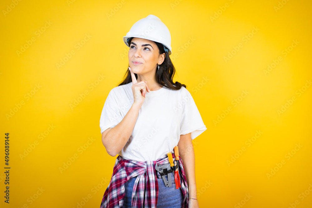 Young caucasian woman wearing hardhat and builder clothes over isolated yellow background happy face thinking about question with hand on chin, thoughtful about confusing idea Young caucasian woman wearing hardhat and builder clothes over isolated yellow background happy face thinking about question with hand on chin, thoughtful about confusing idea