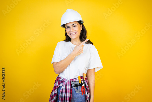 Young caucasian woman wearing hardhat and builder clothes over isolated yellow background smiling and pointing with hand and finger to the side