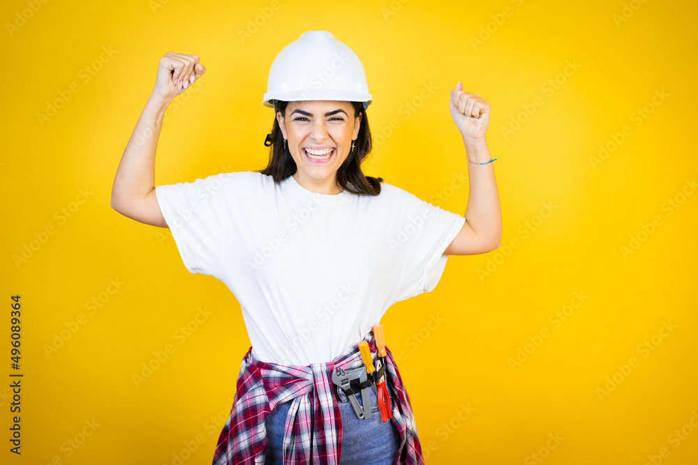 Young caucasian woman wearing hardhat and builder clothes over isolated yellow background very happy and excited making winner gesture with raised arms, smiling and screaming for success.