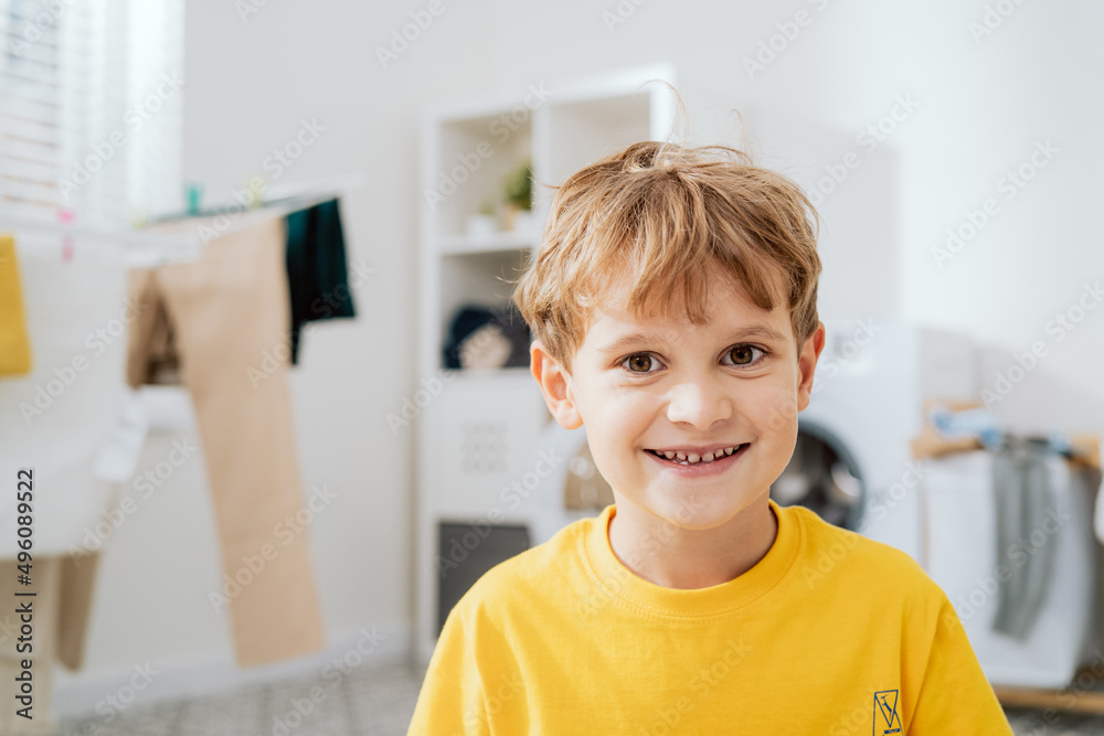 Portrait of smiling child in yellow T-shirt spending time in bathroom laundry room boy helps with household chores putting clothes in washing machine and hanging them on dryer Portrait of smiling child in yellow T-shirt spending time in bathroom laundry room boy helps with household chores putting clothes in washing machine and hanging them on dryer
