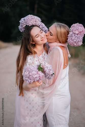 Mom kisses her daughter on the cheek. Two cute girls in white dresses hugging outdoors holding flowers in their hands. Smiling girl with long dark hair. Family photo of mother and daughter. photo