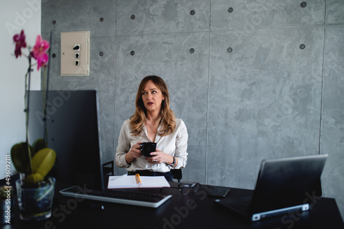 Serious face caucasian middle-aged woman has an essential dialogue with her client/employee.Holds black mug of tea/coffee with both hands.Modern gray wall behind her.Glasses,notebook,equipment, pen.