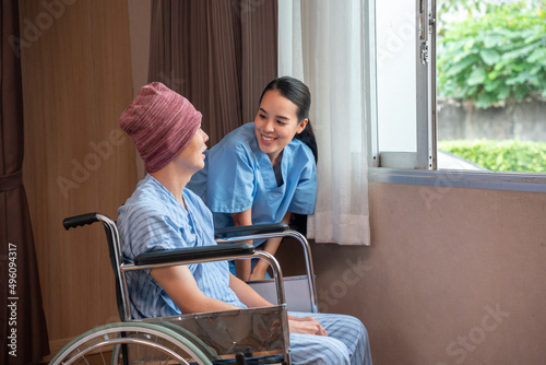 A smiling young Asian female nurse having fun talking with an Asian male cancer patient in a head cover sitting on a wheelchair, while taking him to relax with green nature outside the open window.