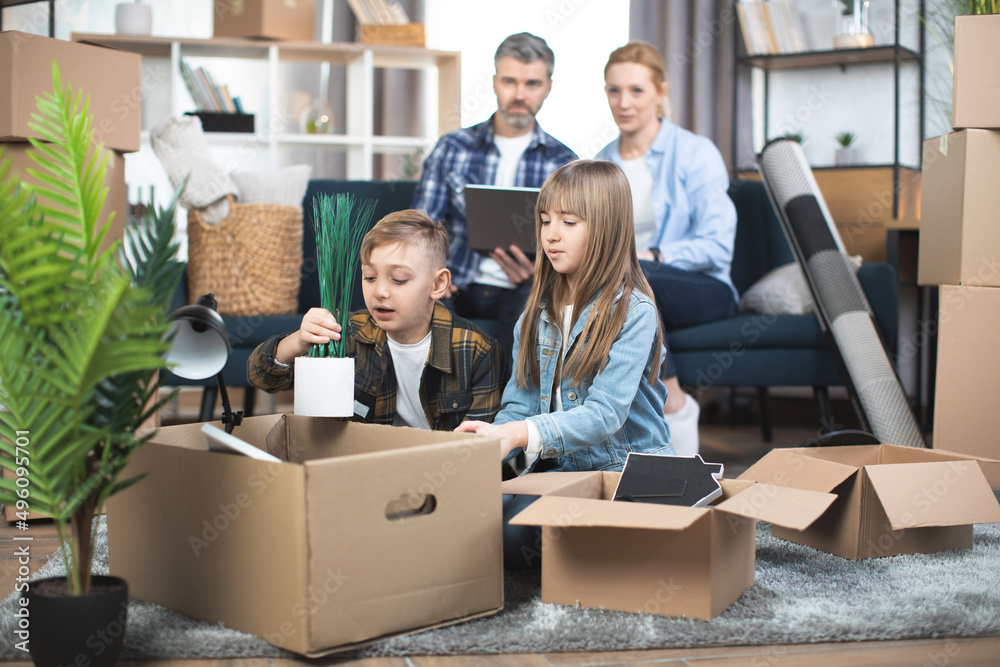 Caucasian couple sitting on couch and using laptop, while their excited kids, boy and girl, unpacking moving boxes and playing together on floor. Concept of relocation and mortgage. Caucasian couple sitting on couch and using laptop, while their excited kids, boy and girl, unpacking moving boxes and playing together on floor. Concept of relocation and mortgage.