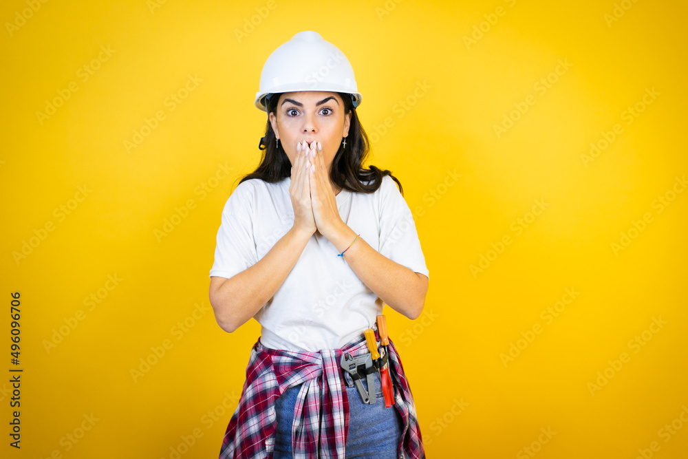 Young caucasian woman wearing hardhat and builder clothes over isolated yellow background shocked covering mouth with hands for mistake. secret concept.
