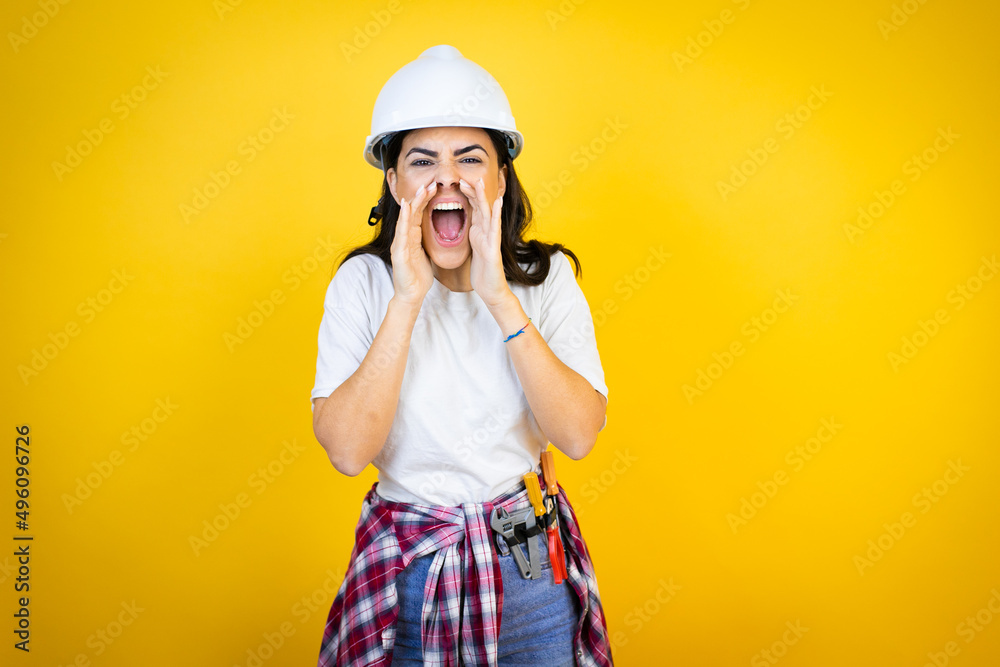 Young caucasian woman wearing hardhat and builder clothes over isolated yellow background shouting and screaming loud to side with hands on mouth Young caucasian woman wearing hardhat and builder clothes over isolated yellow background shouting and screaming loud to side with hands on mouth