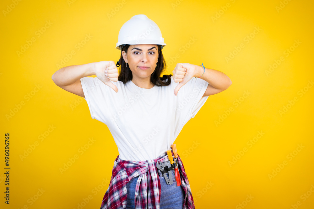 Young caucasian woman wearing hardhat and builder clothes over isolated yellow background with angry face, negative sign showing dislike with thumbs down Young caucasian woman wearing hardhat and builder clothes over isolated yellow background with angry face, negative sign showing dislike with thumbs down
