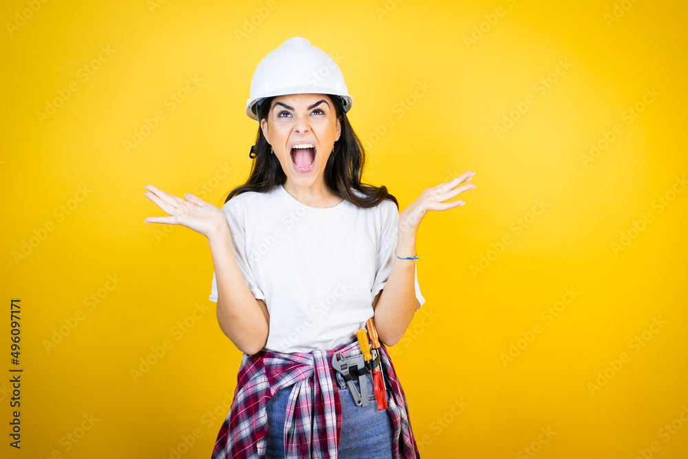 Young caucasian woman wearing hardhat and builder clothes over isolated yellow background clueless and confused expression with arms and hands raised