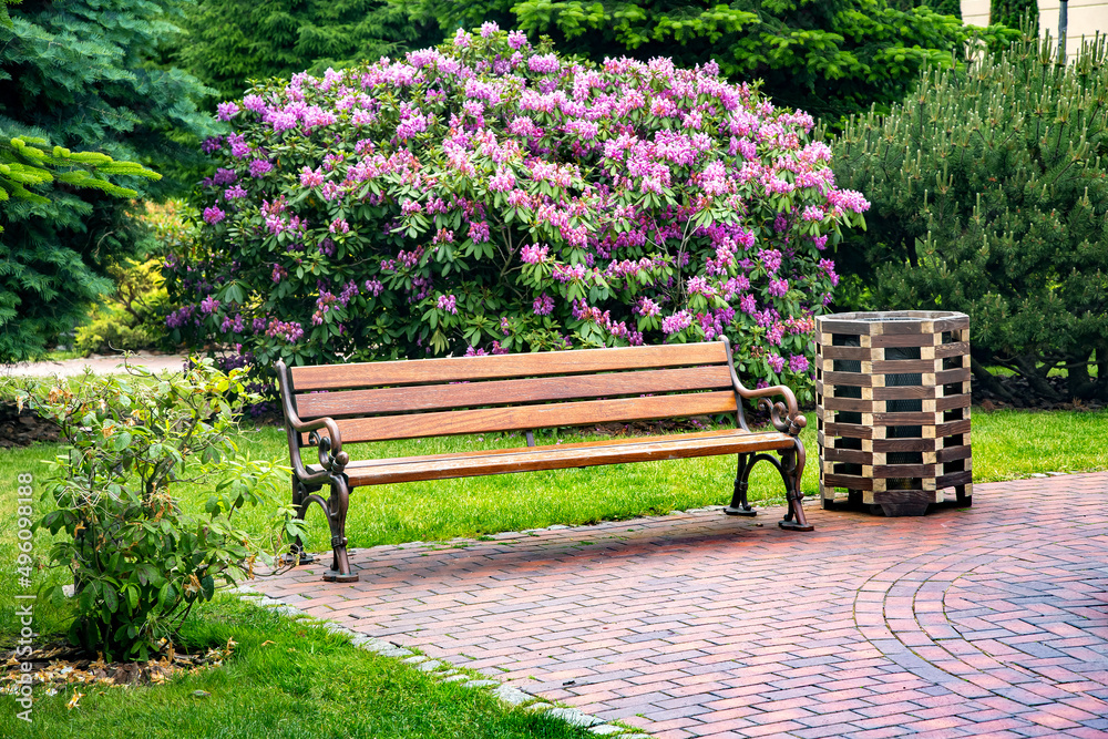 wooden bench with iron legs with a waste bin made of planks on stone tiles in a garden with green plants and flowering bushes, backyard spring landscape empty seat nobody. wooden bench with iron legs with a waste bin made of planks on stone tiles in a garden with green plants and flowering bushes, backyard spring landscape empty seat nobody.