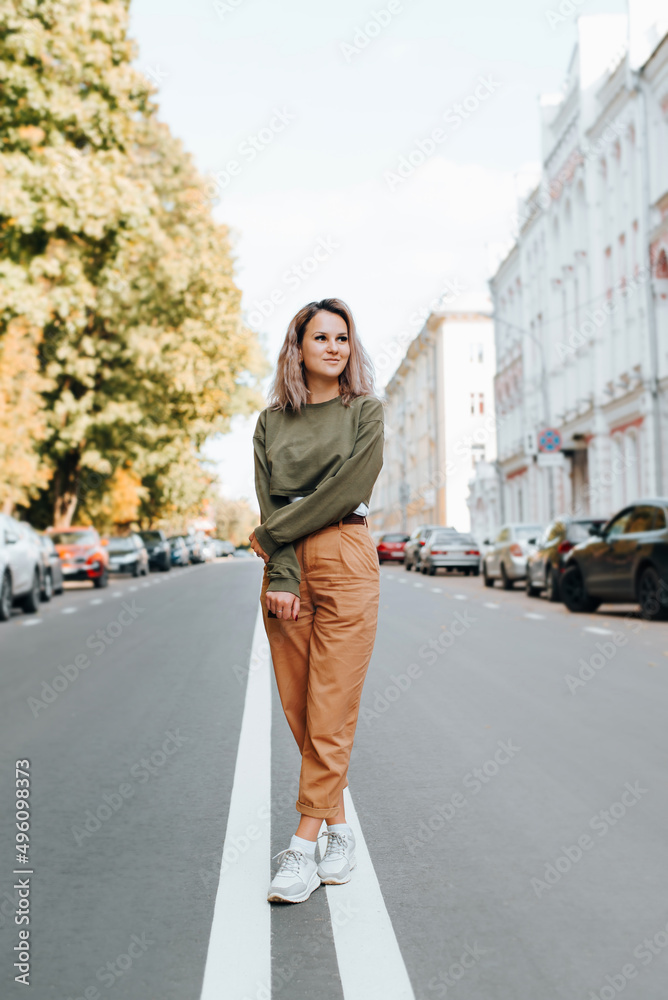 Hipster in youthful casual clothes with cute smile standing on road markings on street and looking away. Pretty young caucasian woman posing in city during day outdoors