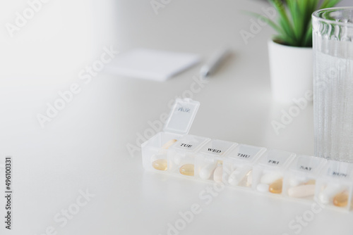 Closeup of medical pill box with doses of tablets for daily take a medicine with white, yellow drugs and capsules used for treatment, cure the disease. Glass cup of water on table, gray background.