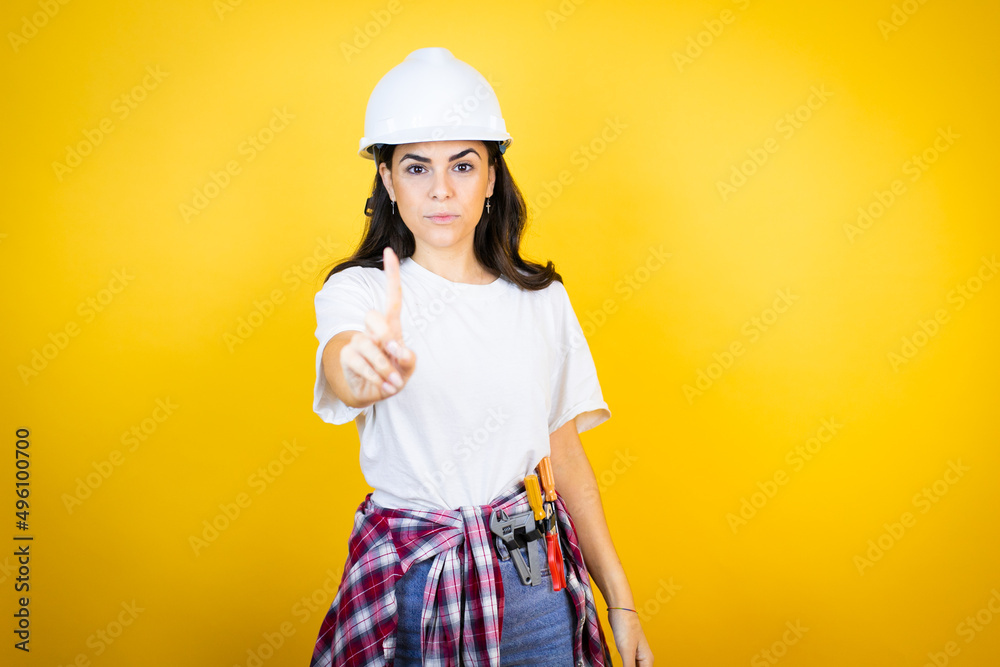 Young caucasian woman wearing hardhat and builder clothes over isolated yellow background showing and pointing up with fingers number one while is serious Young caucasian woman wearing hardhat and builder clothes over isolated yellow background showing and pointing up with fingers number one while is serious