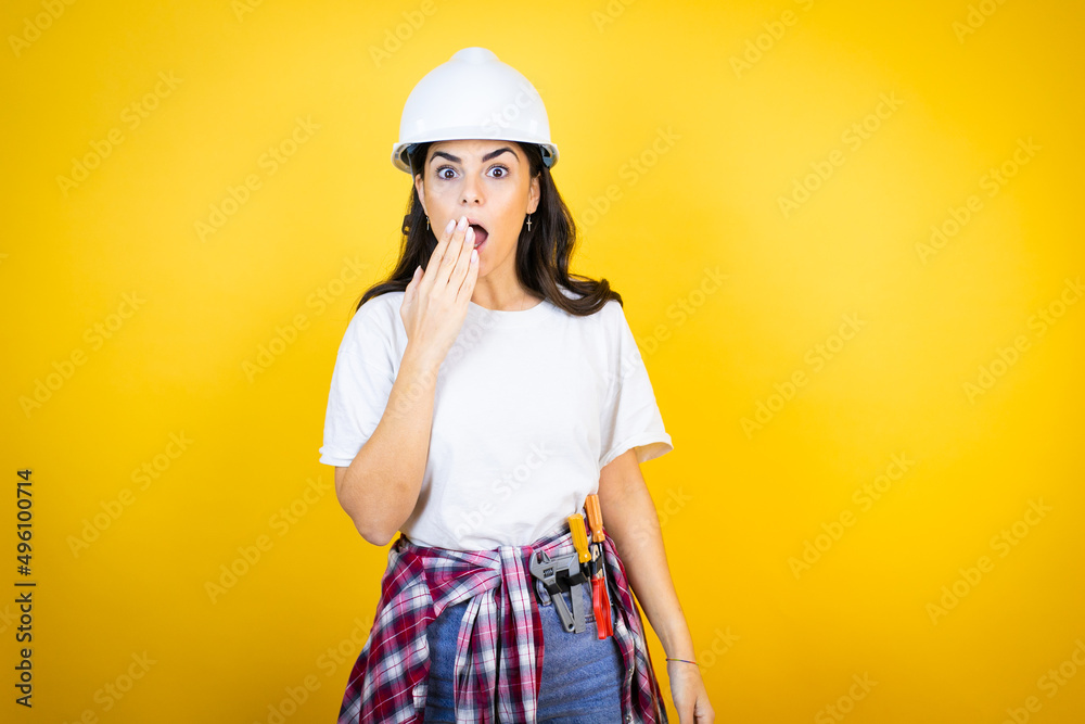 Young caucasian woman wearing hardhat and builder clothes over isolated yellow background covering mouth with hand, shocked and afraid for mistake. surprised expression Young caucasian woman wearing hardhat and builder clothes over isolated yellow background covering mouth with hand, shocked and afraid for mistake. surprised expression