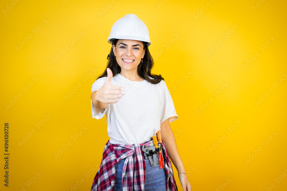 Young caucasian woman wearing hardhat and builder clothes over isolated yellow background smiling friendly offering handshake as greeting and welcoming Young caucasian woman wearing hardhat and builder clothes over isolated yellow background smiling friendly offering handshake as greeting and welcoming