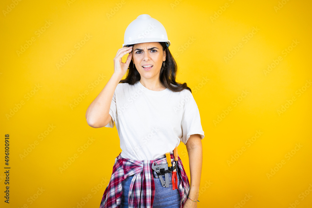 Young caucasian woman wearing hardhat and builder clothes over isolated yellow background confuse and wonder about question. Uncertain with doubt, thinking with hand on head Young caucasian woman wearing hardhat and builder clothes over isolated yellow background confuse and wonder about question. Uncertain with doubt, thinking with hand on head