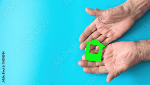 The hands of an elderly man hold a house cut out of paper on a blue background. ecological house. Save nature and ecology. Nursing home theme. Lonely old age., homeless person. Copyspace