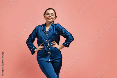 Portrait of young smiling woman in blue silk pajamas posing isolated over pink studio background