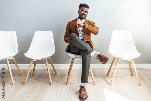 Full length shot of a handsome young businessman sitting and looking down at his watch against a gray studio background. I'm getting bored waiting for them photo