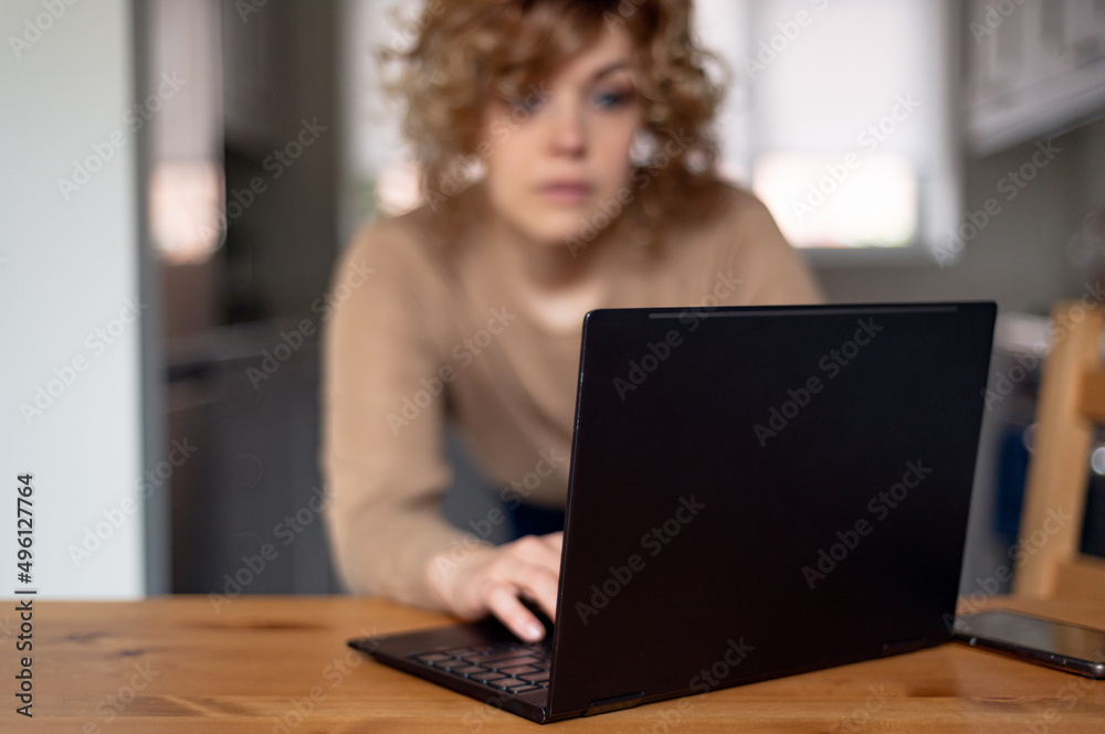 Blurred Portrait of serious woman who is looking at laptop in her kitchen, working from home. Using computer for video call, looking at screen or reading news Blurred Portrait of serious woman who is looking at laptop in her kitchen, working from home. Using computer for video call, looking at screen or reading news