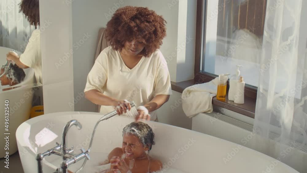 High angle shot of careful African American mother rinsing shampoo out of hair of little son while washing kid in bathroom at home