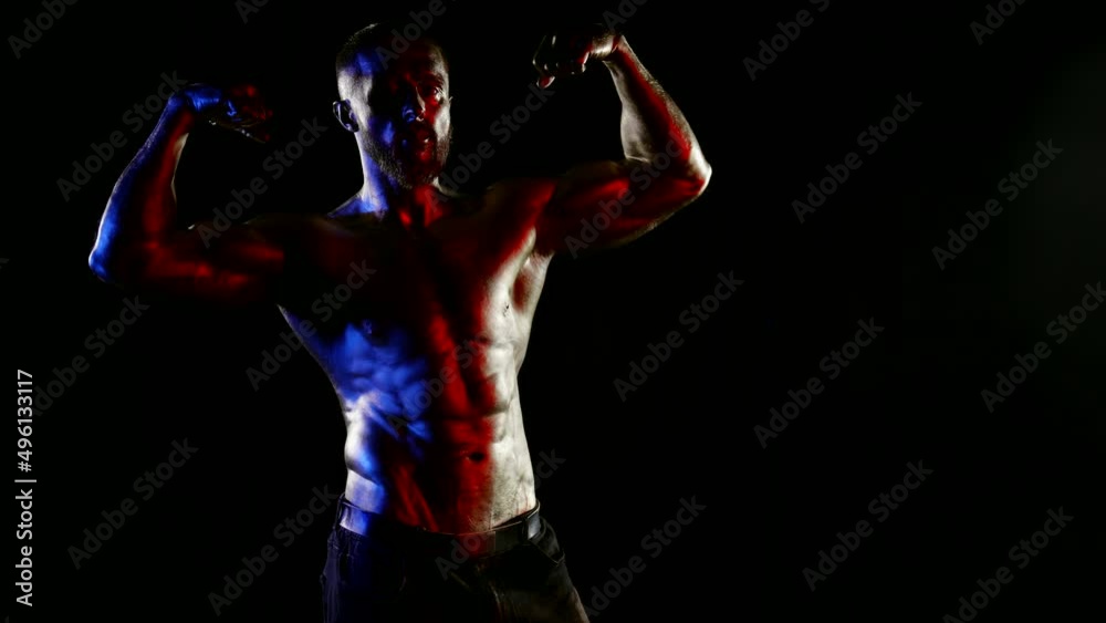 a man with a muscular naked torso and metallic golden skin poses and demonstrates biceps on a black background. red, blue and white light. dark key a man with a muscular naked torso and metallic golden skin poses and demonstrates biceps on a black background. red, blue and white light. dark key