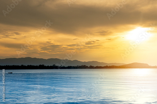 Abstract of wonderful light of the golden sky and the blue sea. with the silhouette of the forests and mountains on the distant shores. Sun shines out from the sky above.
