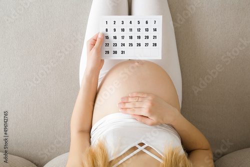Young adult pregnant woman with big naked belly sitting on sofa at home. Hand holding calendar and thinking about childbirth date. Baby expecting concept. Pregnancy time. Closeup. Top down view.