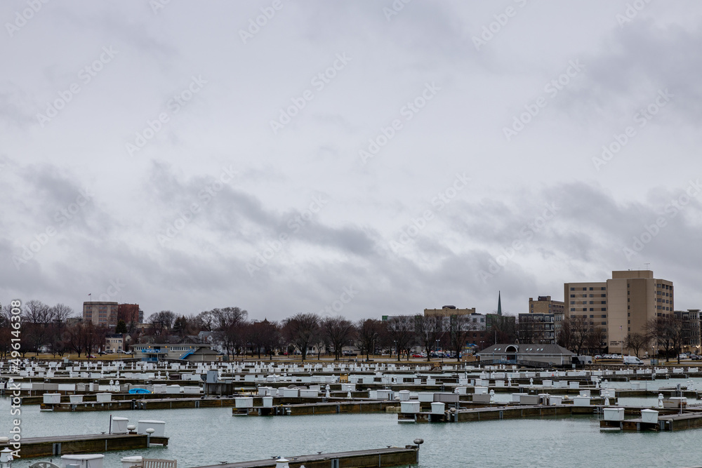 Heavily overcast sky with empty boat marina in autumn. Wood piers and docks with benches and storage containers each. Park and bare trees in background. Marina store building. 