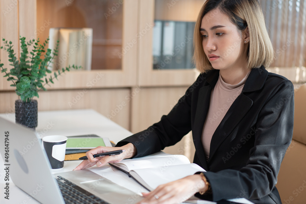 Asian Female manager working on project using computer laptop. reading project report document. work review at desk. checking financial account in the office Asian Female manager working on project using computer laptop. reading project report document. work review at desk. checking financial account in the office