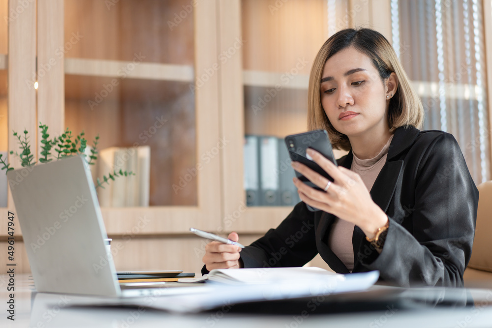 Asian female manager or employee watching mobile phone and working with computer laptop at desk in the office. checking financial account report