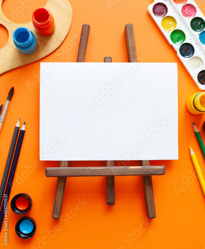 white blank sheet, wooden easel and various art supplies on orange background. copy space. place for text