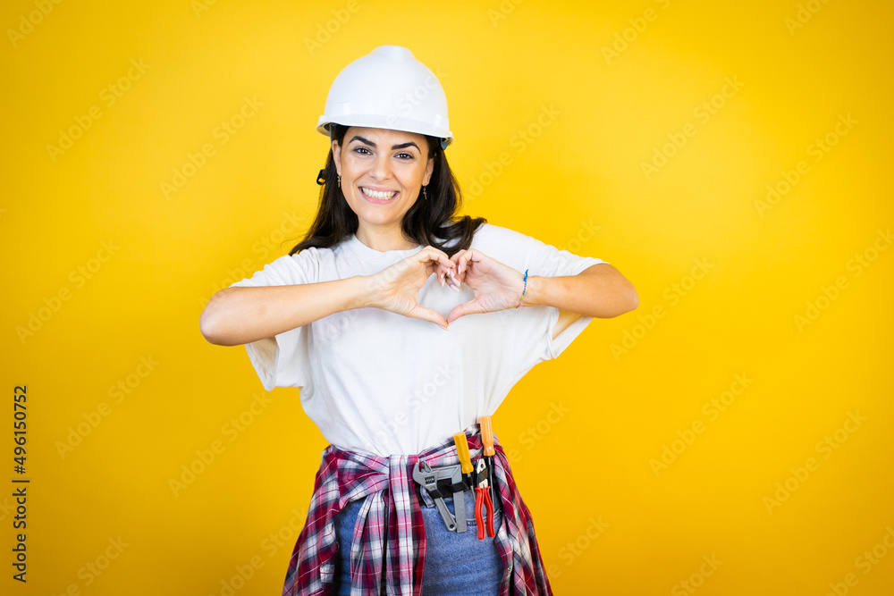 Young caucasian woman wearing hardhat and builder clothes over isolated yellow background smiling in love showing heart symbol and shape with hands Young caucasian woman wearing hardhat and builder clothes over isolated yellow background smiling in love showing heart symbol and shape with hands
