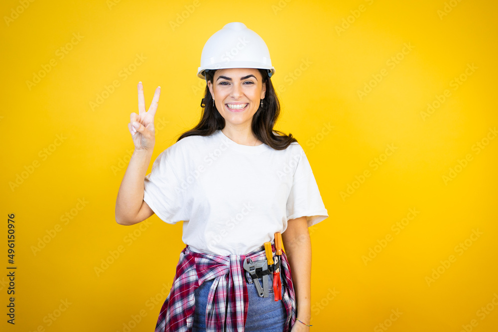 Young caucasian woman wearing hardhat and builder clothes over isolated yellow background showing and pointing up with fingers number two while smiling confident and happy