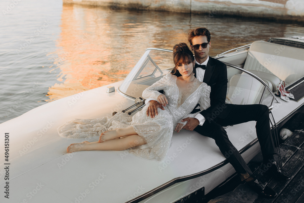 Couple rides a yacht on a sunny day. Stylish groom in a black suit and sunglasses hugs and kisses a bride in wedding dress