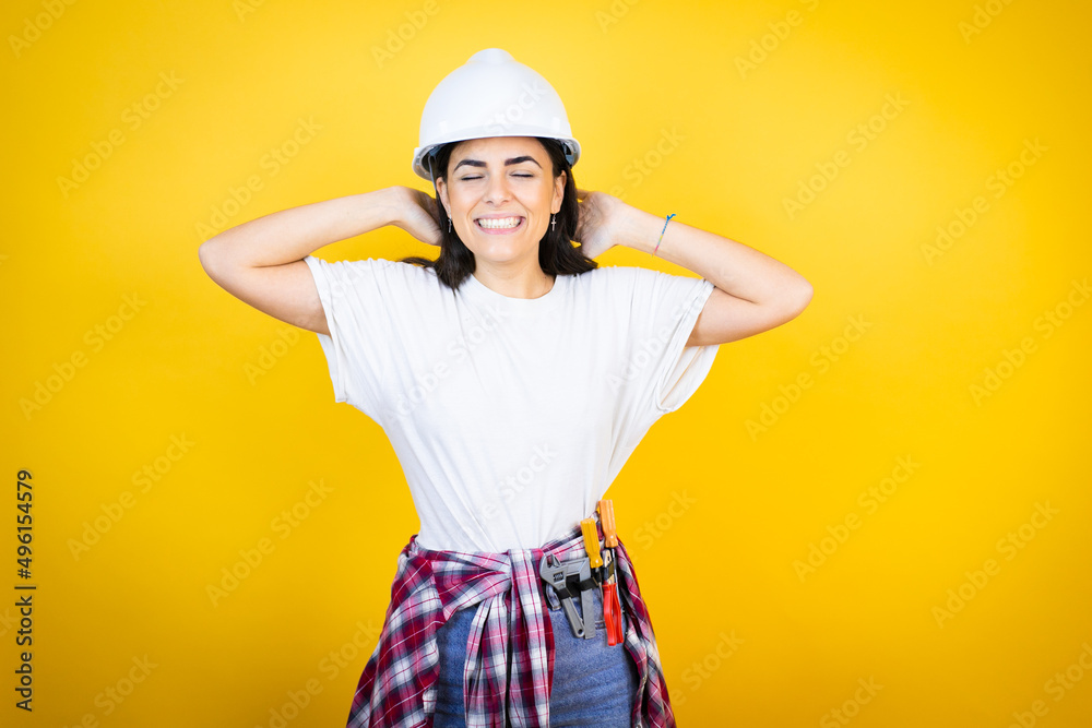 Young caucasian woman wearing hardhat and builder clothes over isolated yellow background relaxing and stretching, arms and hands behind head and neck smiling happy