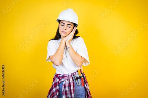 Young caucasian woman wearing hardhat and builder clothes over isolated yellow background sleeping tired dreaming and posing with hands together while smiling with closed eyes. © Irene