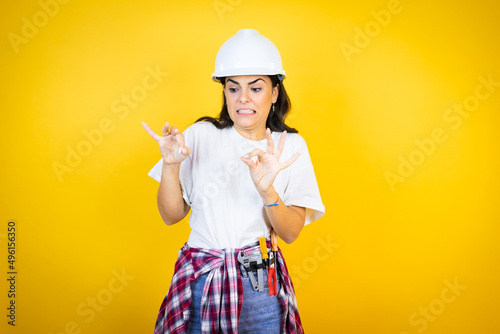 Young caucasian woman wearing hardhat and builder clothes over isolated yellow background disgusted expression, displeased and fearful doing disgust face because aversion reaction.Annoying concept photo